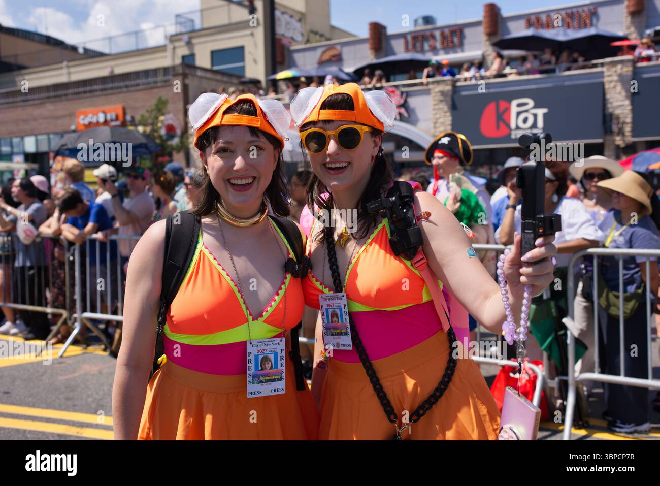 Due donne con costumi da bagno arancione e rosa e cappelli innovativi tengono in mano una macchina fotografica e si mettono in posa per una foto durante un evento all'aperto. Indossano i pass per la stampa. Il ba Foto Stock