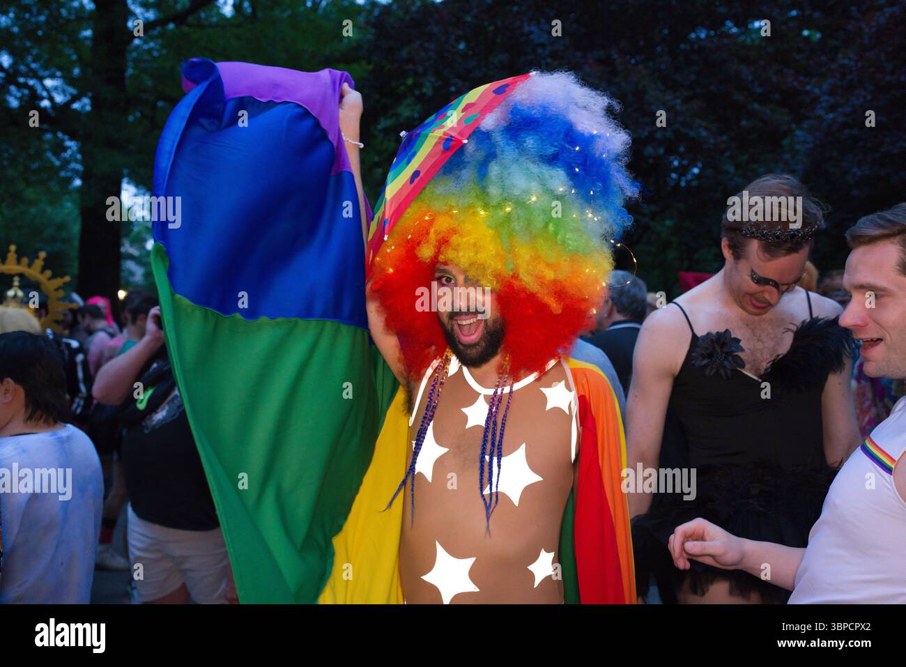 Una persona con una parrucca arcobaleno e un mantello multicolore ha una bandiera di colore simile a un evento Pride. Altre persone in vari costumi sono visibili in Foto Stock