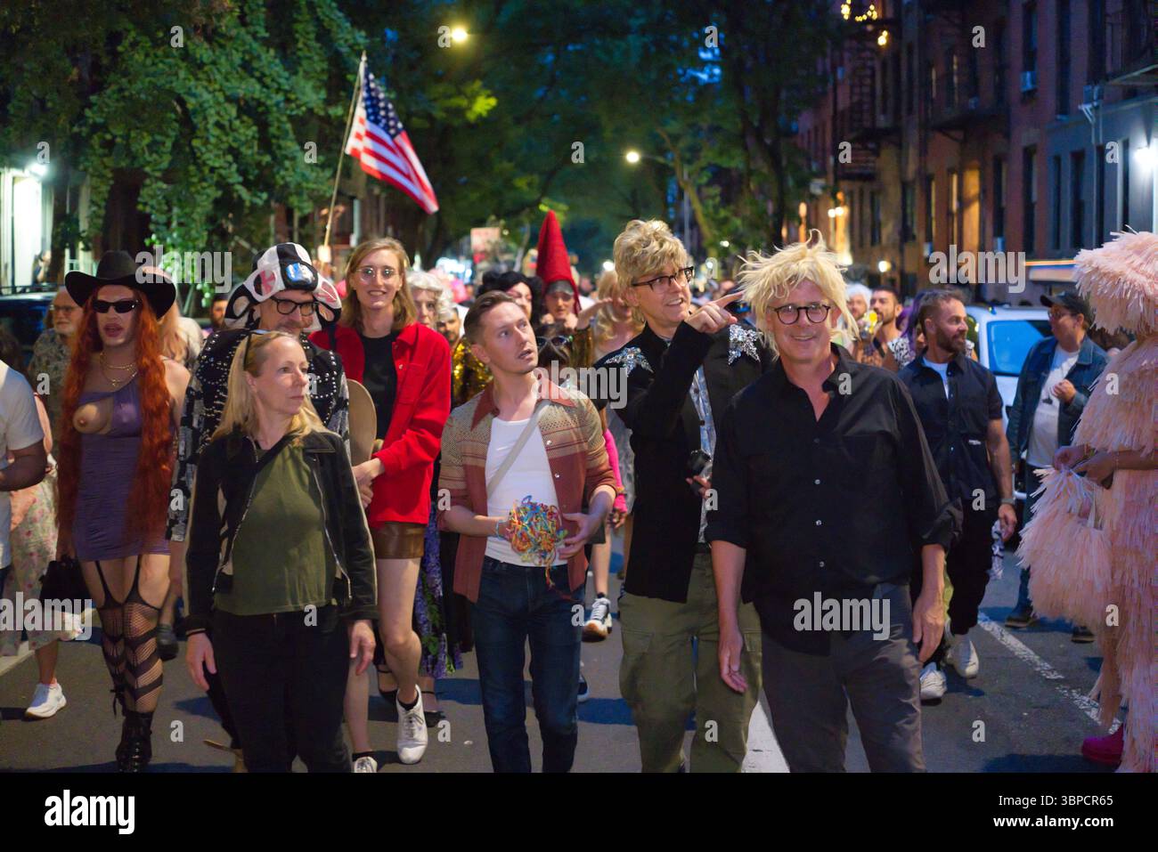 Scena notturna di strada di un gruppo di persone, molte in costume o con un abbigliamento sgargiante, camminando in parata o in processione. Il gruppo comprende individui Foto Stock