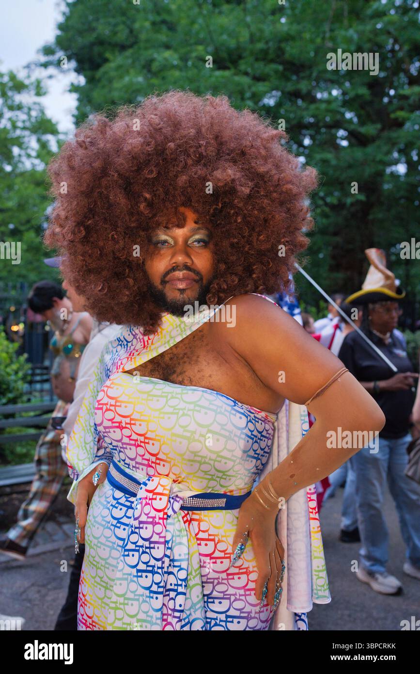 Una persona con un grande afro marrone indossa un abito con motivo arcobaleno, un abito a spalla e guanti lunghi. Stanno posando all'aperto, parzialmente visibili ad altri Foto Stock