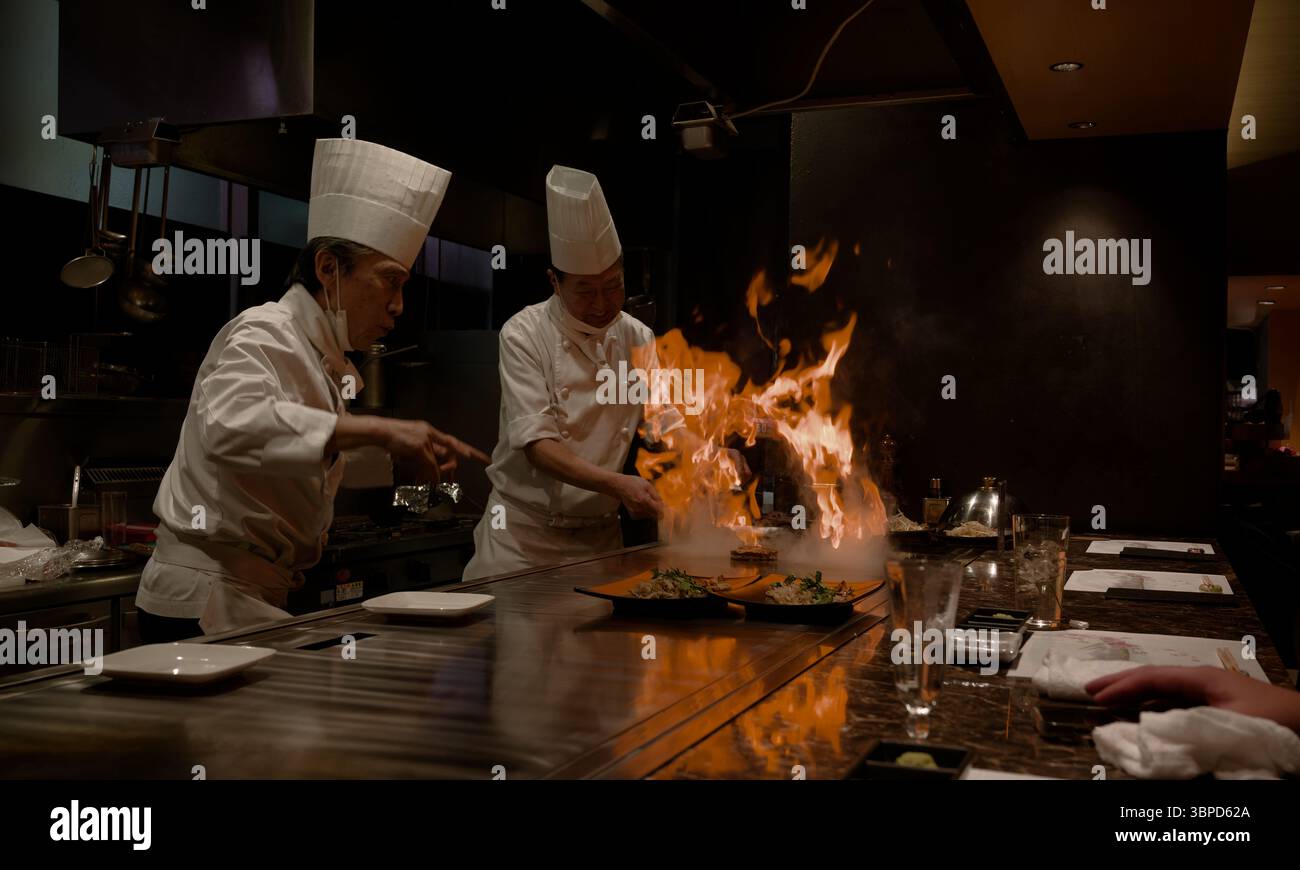 Gli chef Teppanyaki a Tokyo creano un drammatico scoppio di fiamme sulla griglia mentre i commensali guardano, mostrando abilità e talento in un ambiente vivace Foto Stock