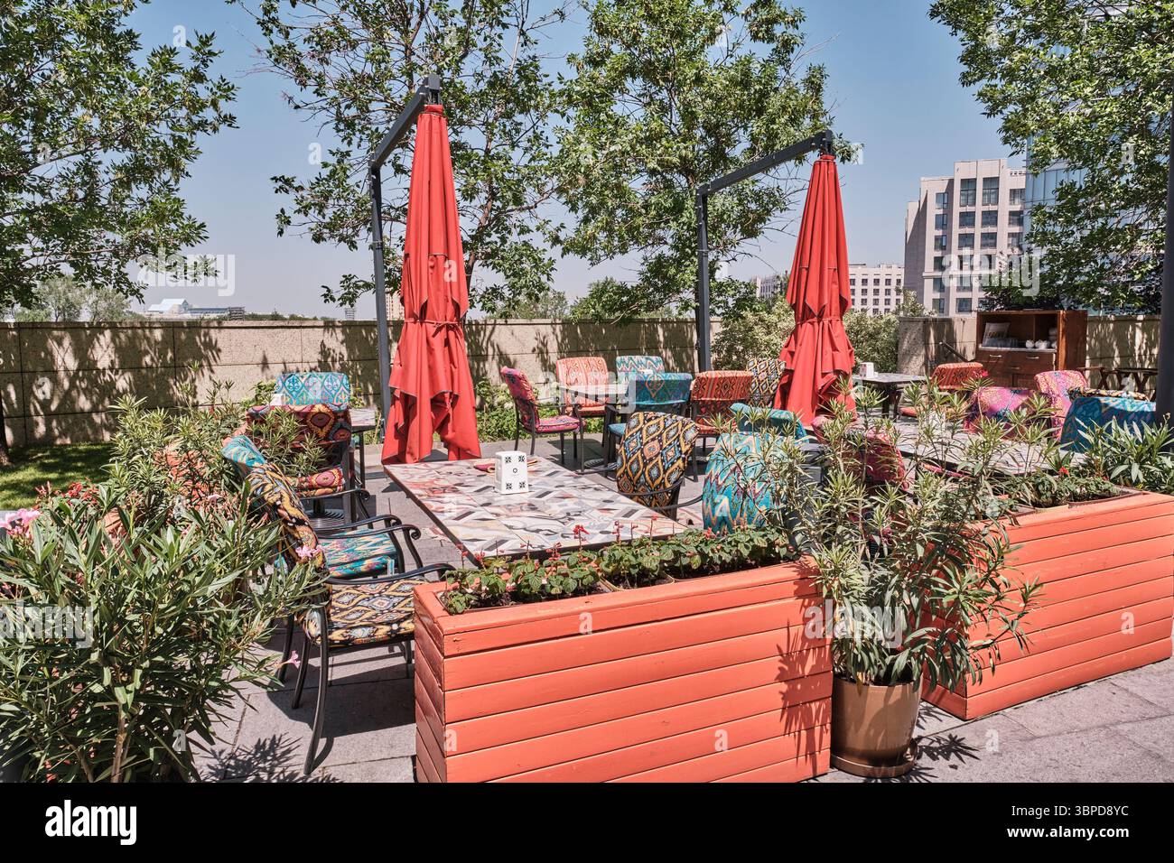 Kazakistan, Almaty - 28 giugno 2025: Vivace terrazza all'aperto del ristorante con sedie a motivi geometrici, ombrelli rossi e decorazioni in stile orientale, circondata da Foto Stock