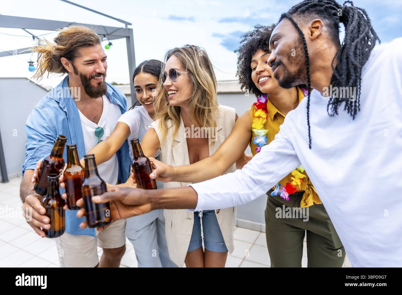 Amici felici che festeggiano sul tetto, brindano con bottiglie di birra e si godono momenti di qualità insieme in una vibrante atmosfera estiva Foto Stock