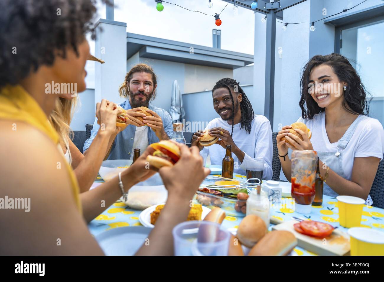 Allegri amici multietnici gustando hamburger e bevande mentre vi godete una vivace festa al barbecue sul tetto piena di risate e connessioni Foto Stock