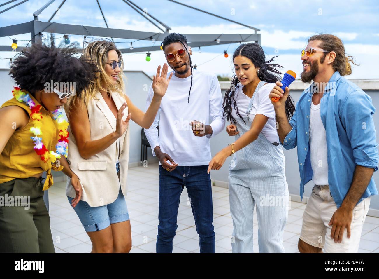Gruppo di allegri amici multietnici che si divertono a cantare karaoke e ballare insieme durante una festa sul tetto Foto Stock