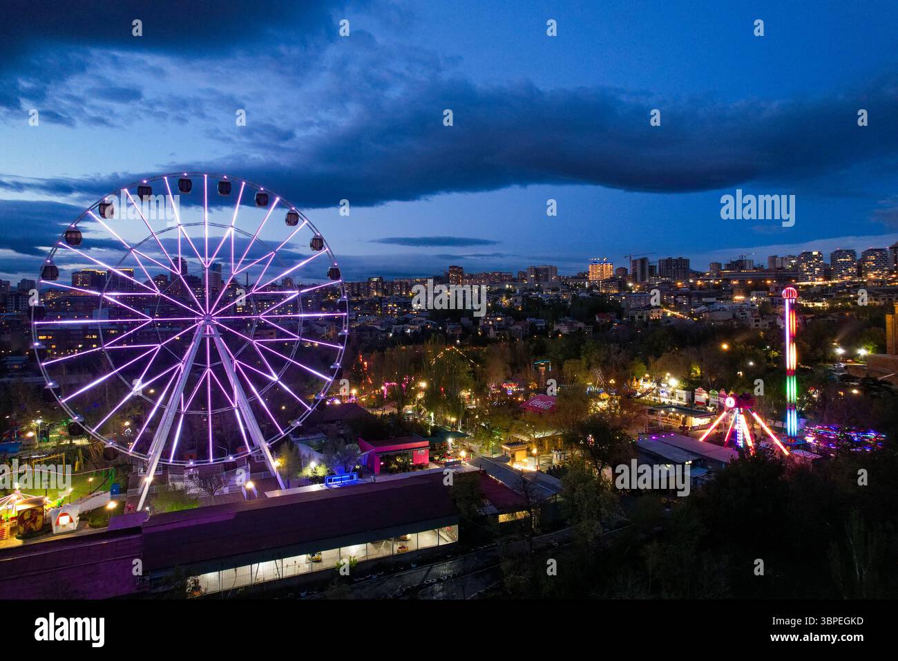 Una ruota panoramica illuminata da luci viola al crepuscolo nel Parco della Vittoria, Erevan, Armenia, che si affaccia sullo skyline della città e sulle attrazioni del parco divertimenti. Foto Stock