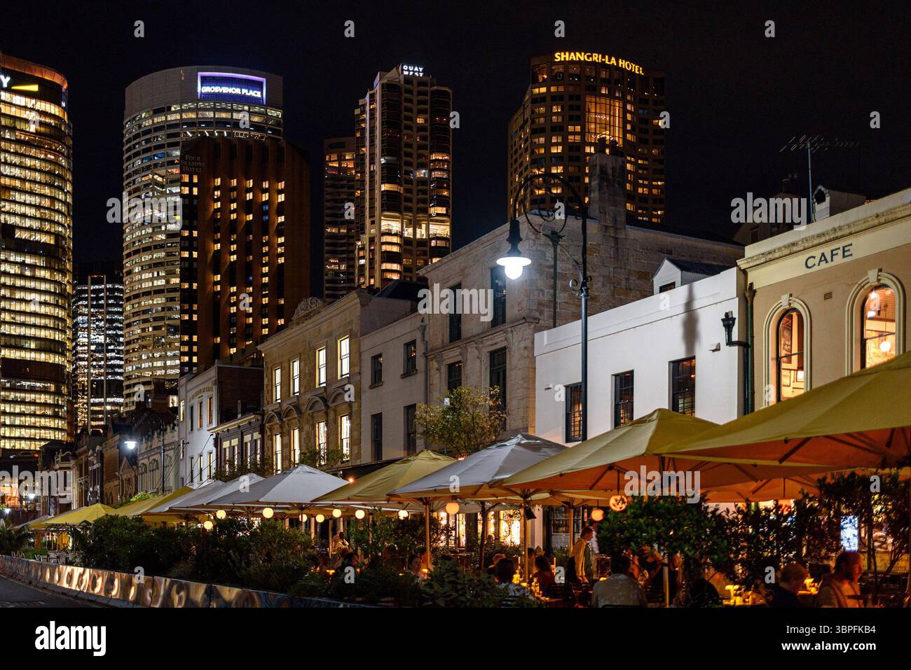 Persone sedute su terrazze lungo George Street di notte nell'area di Rocks di Sydney con grattacieli dal CBD sullo sfondo Foto Stock