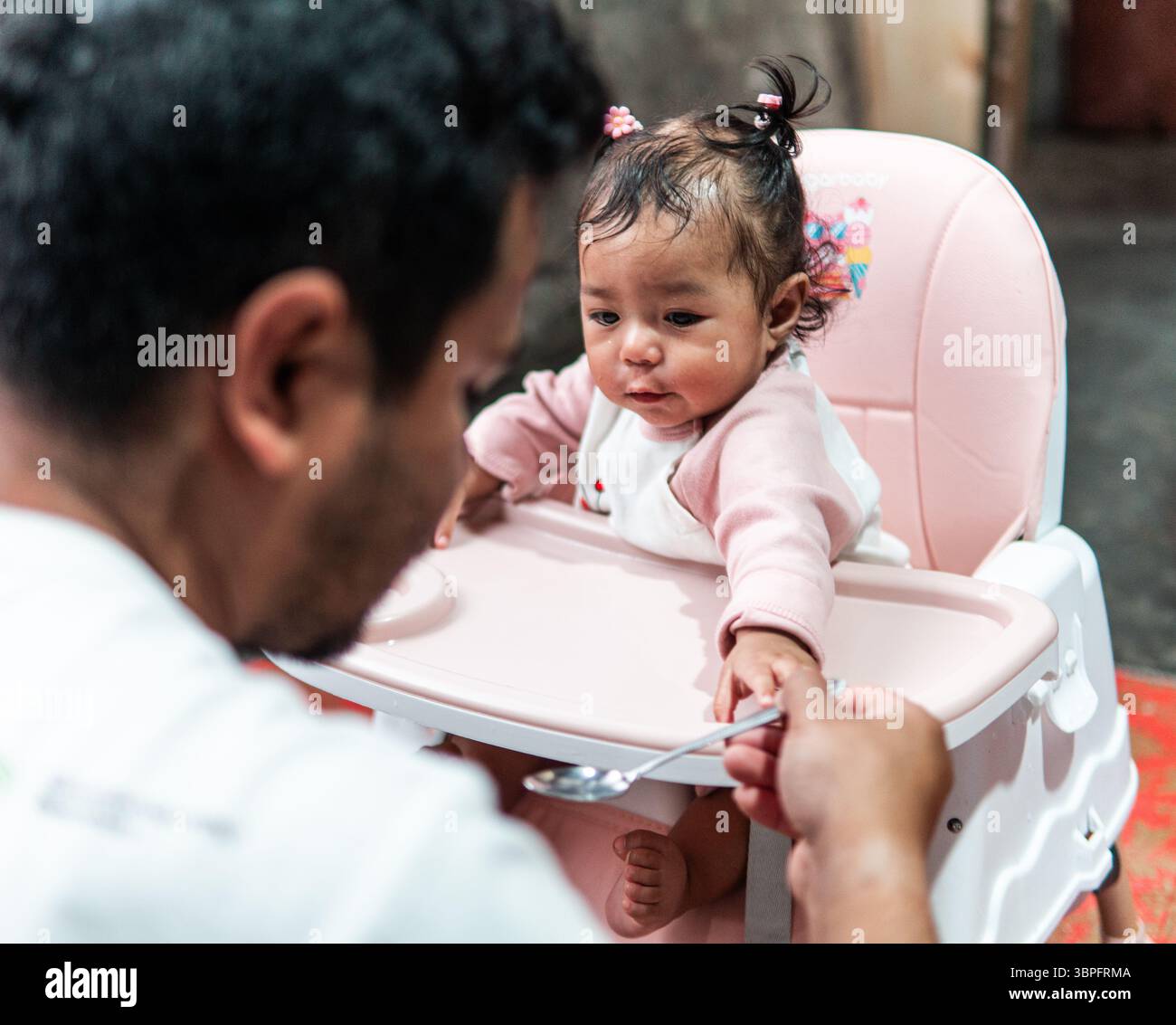 Una bambina carina seduta su un seggiolone sembra curiosa mentre suo padre le dà da mangiare con un cucchiaio. Un emozionante momento di amore e cura per tutta la famiglia Foto Stock