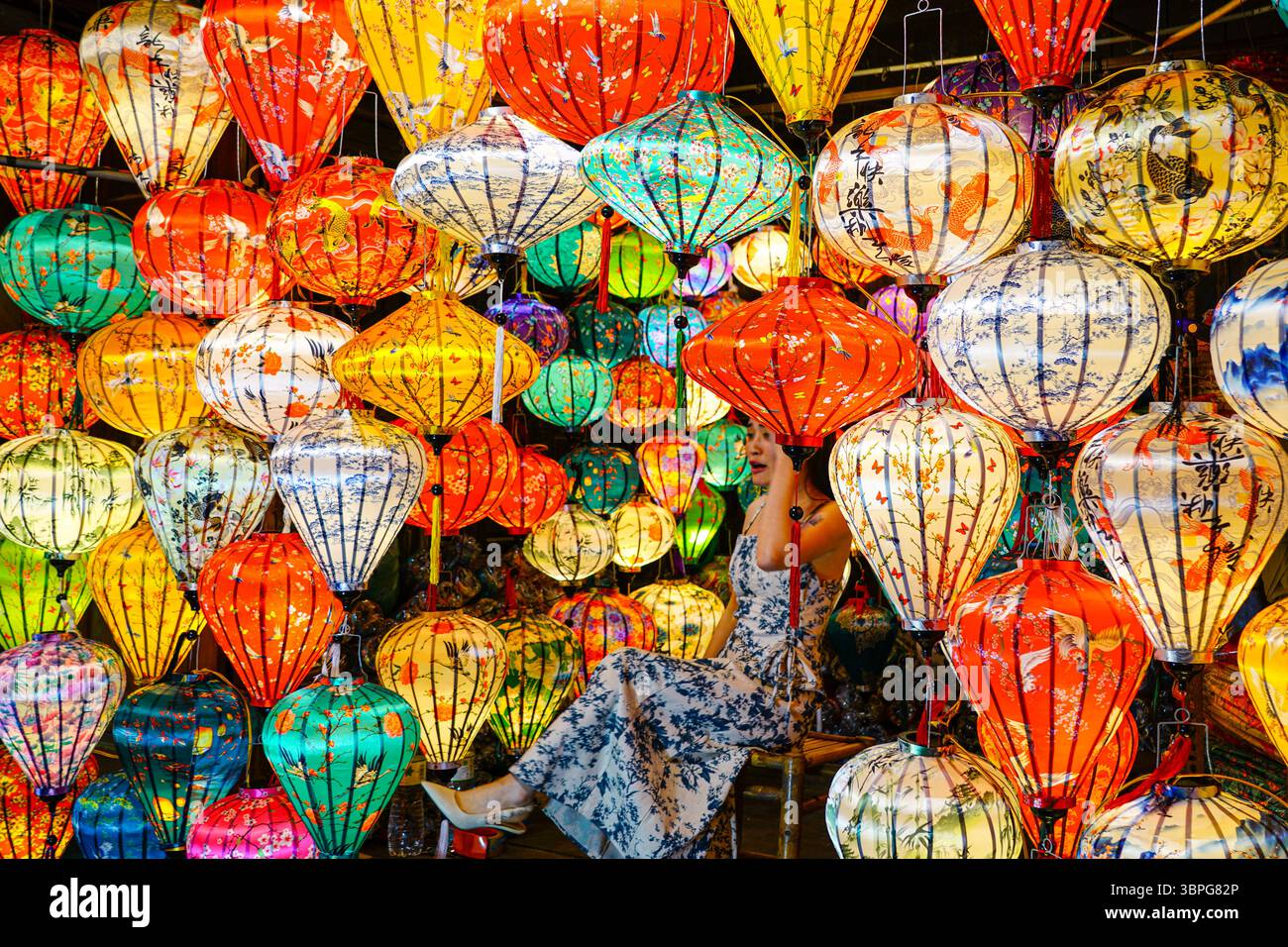 Da Nang, Vietnam - 18 marzo 2025: Donna in abito floreale siede tra lanterne di seta incandescenti, immersa in un mondo scintillante di colori, luci e fascino culturale Foto Stock