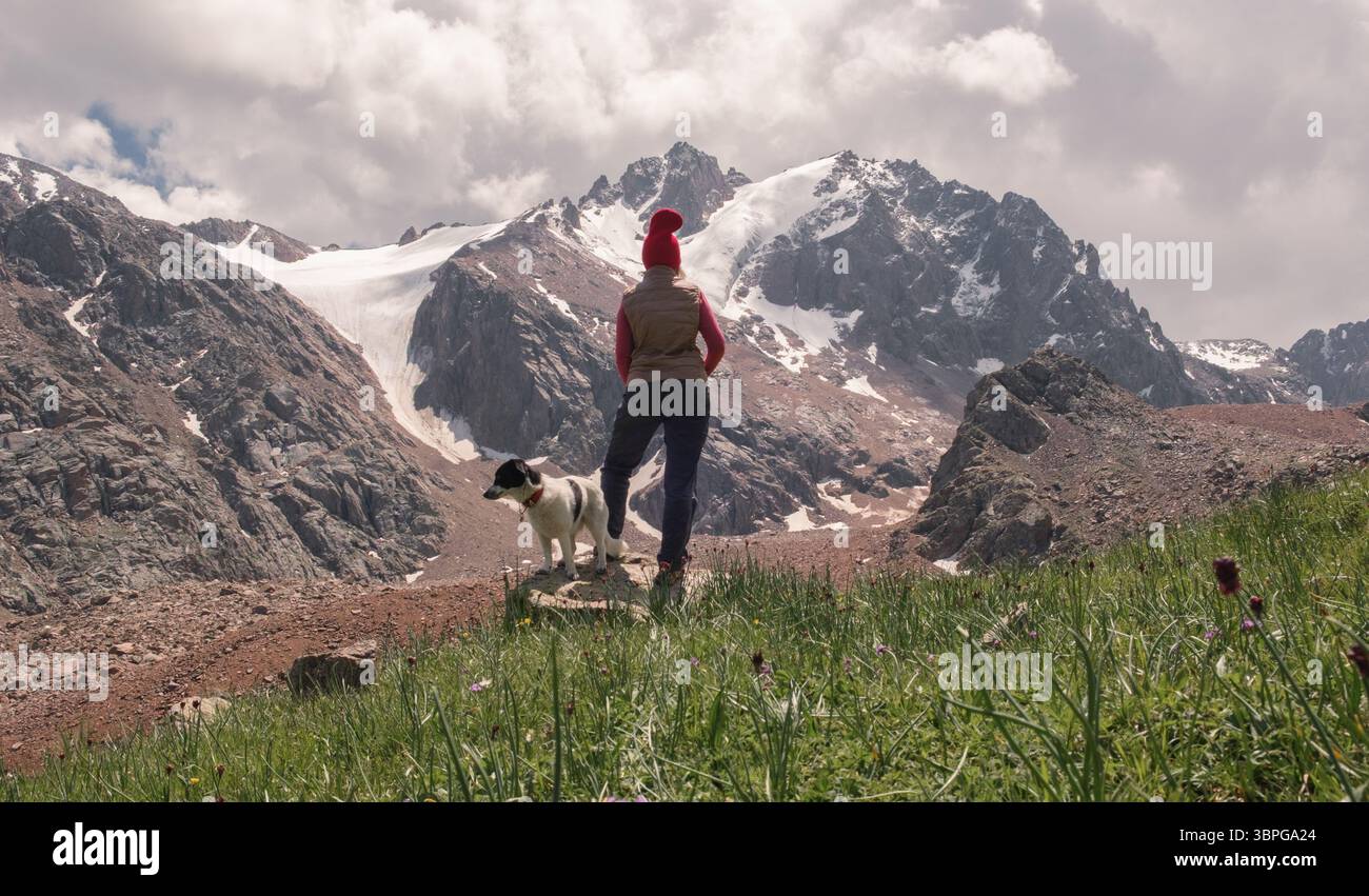 Vista posteriore della donna con abiti da trekking, gilet e cappello rosso sullo sfondo delle montagne, sta camminando con il suo cane guardando impressionanti innevate Foto Stock