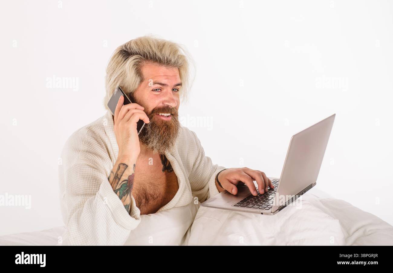 Uomo sorridente barbuto che parla con lo smartphone e digita sul laptop a letto a casa. Stile di vita digitale, multitasking moderno. Bell'uomo che lavora a casa si' Foto Stock