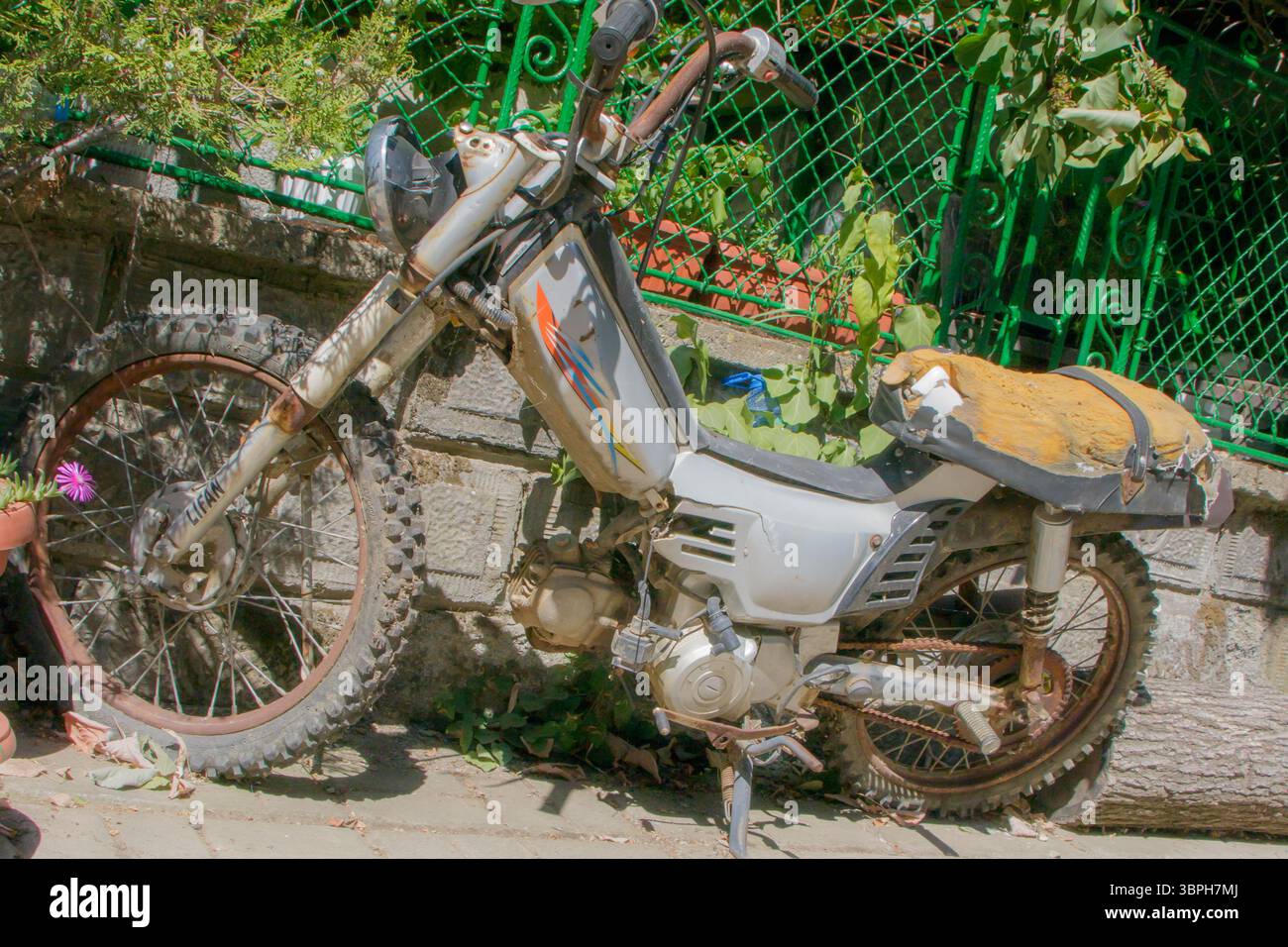 Una motocicletta resistente agli agenti atmosferici si appoggia contro un muro di pietra tra piante verdi. Gli pneumatici sono polverosi e il sedile presenta una fodera del sedile usurata. La luce del sole proietta in gent Foto Stock