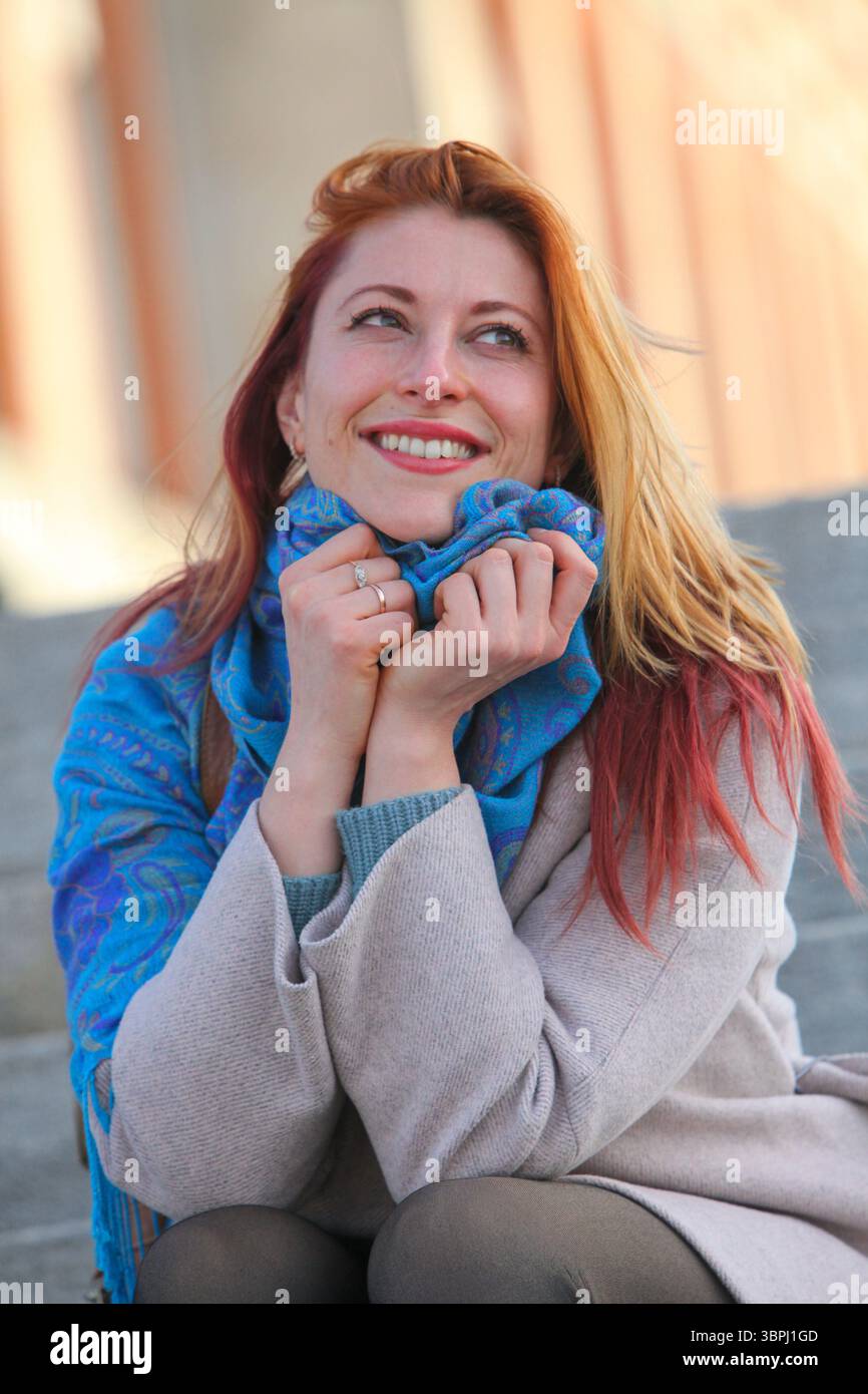 Donna dai capelli di zenzero, anni '30, seduta su scale all'aperto, allegra ed elegante con cappotto e sciarpa. Ritratto autunnale o primaverile con luce naturale. Foto Stock