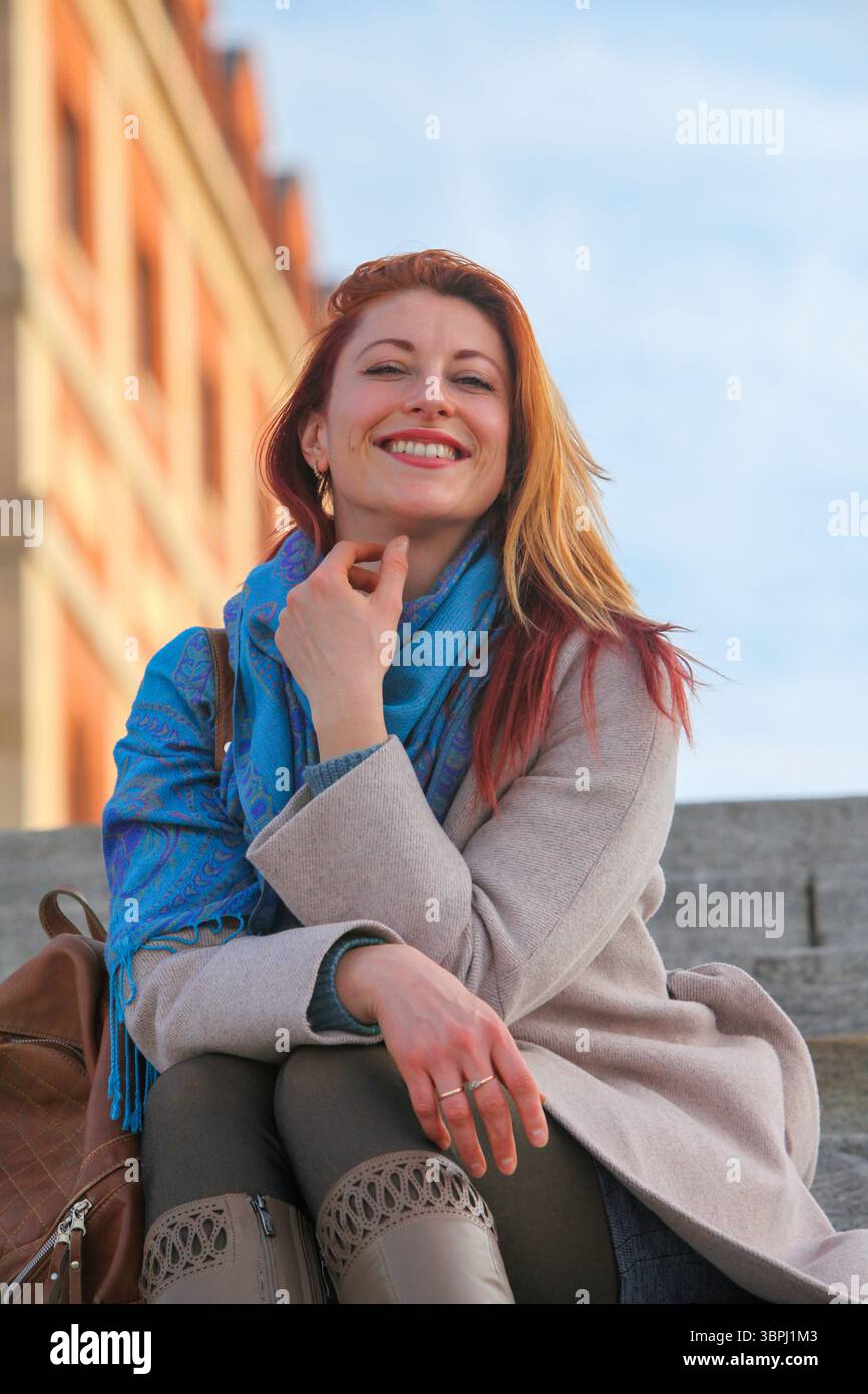 Donna dai capelli di zenzero, anni '30, seduta su scale all'aperto, allegra ed elegante con cappotto e sciarpa. Ritratto autunnale o primaverile con luce naturale. Foto Stock