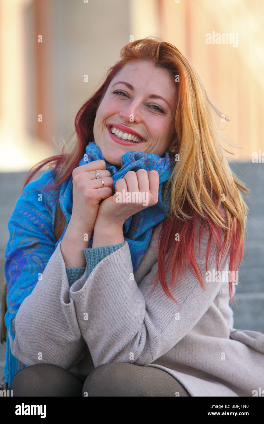 Donna dai capelli di zenzero, anni '30, seduta su scale all'aperto, allegra ed elegante con cappotto e sciarpa. Ritratto autunnale o primaverile con luce naturale. Foto Stock