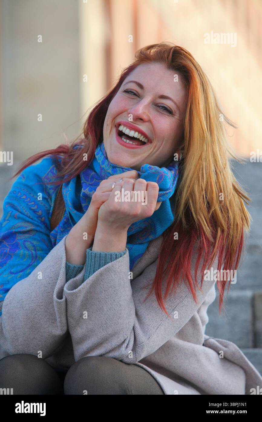 Donna dai capelli di zenzero, anni '30, seduta su scale all'aperto, allegra ed elegante con cappotto e sciarpa. Ritratto autunnale o primaverile con luce naturale. Foto Stock