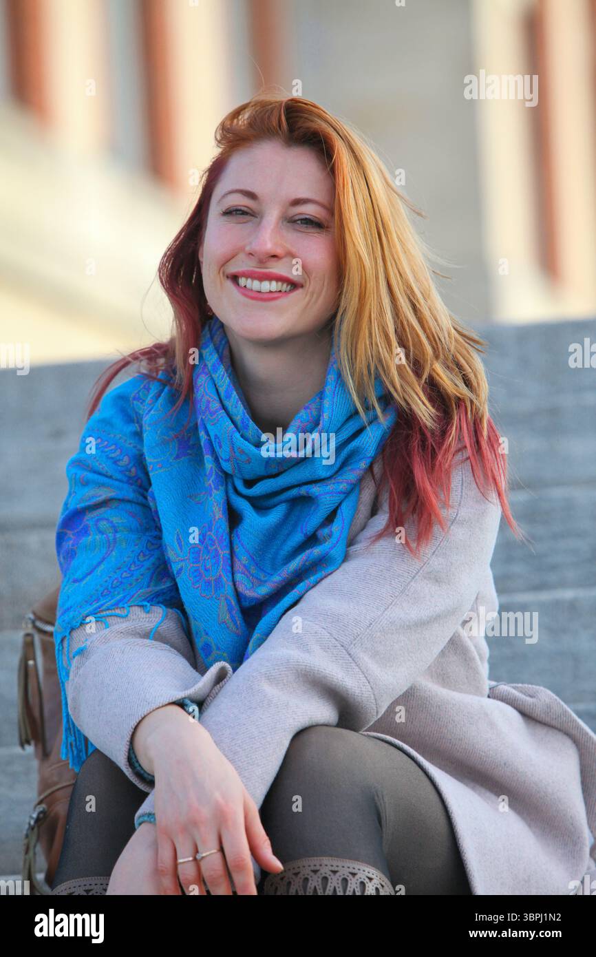 Donna dai capelli di zenzero, anni '30, seduta su scale all'aperto, allegra ed elegante con cappotto e sciarpa. Ritratto autunnale o primaverile con luce naturale. Foto Stock