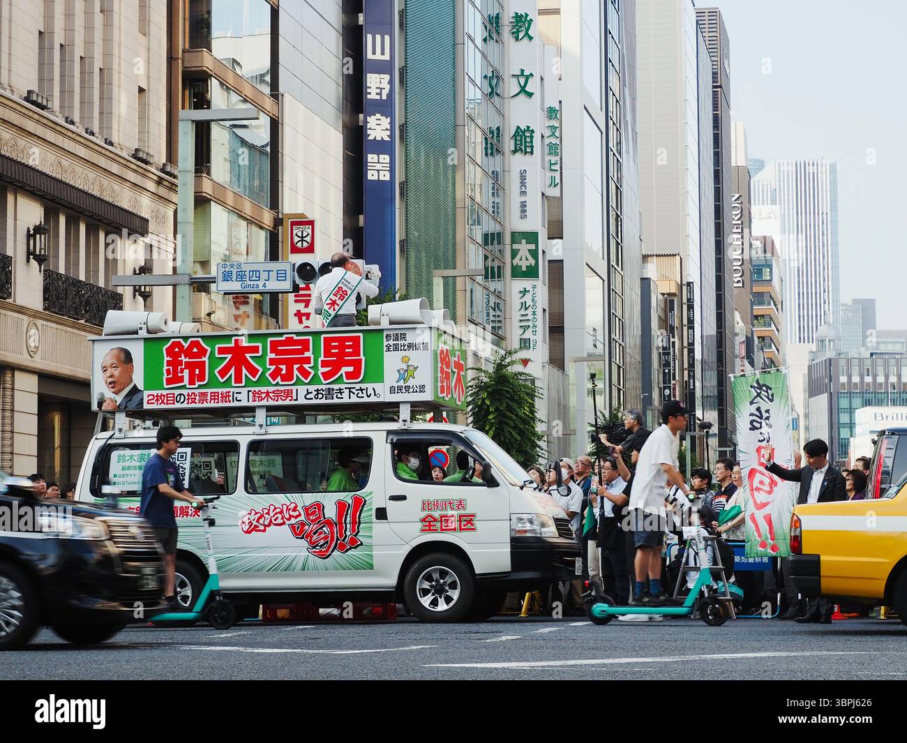 Il politico Muneo Suzuki tiene un discorso a Ginza a Tokyo durante la campagna elettorale della camera dei consiglieri con persone che guidano Luup noleggio e-scooter di passaggio Foto Stock