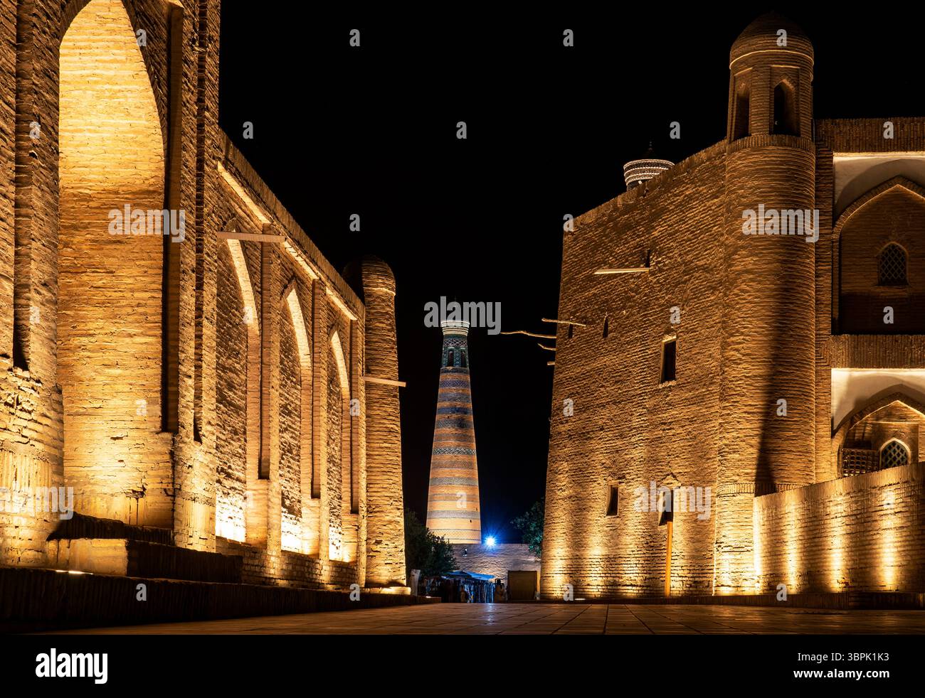 Una vista notturna dell'architettura storica caratterizzata da pareti in mattoni illuminati e un colorato minareto sullo sfondo, che mostra una miscela di antico e. Foto Stock