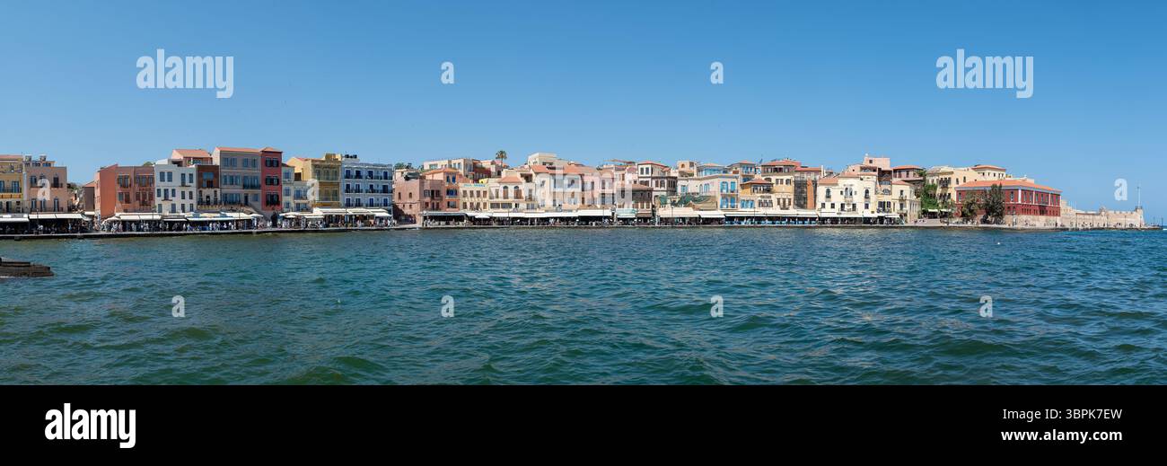 Chania, Creta, Grecia - 22 giugno 2025: Edifici vivaci si riflettono nell'acqua mentre la gente si gode i caffè lungo il porto di Chania. Foto Stock