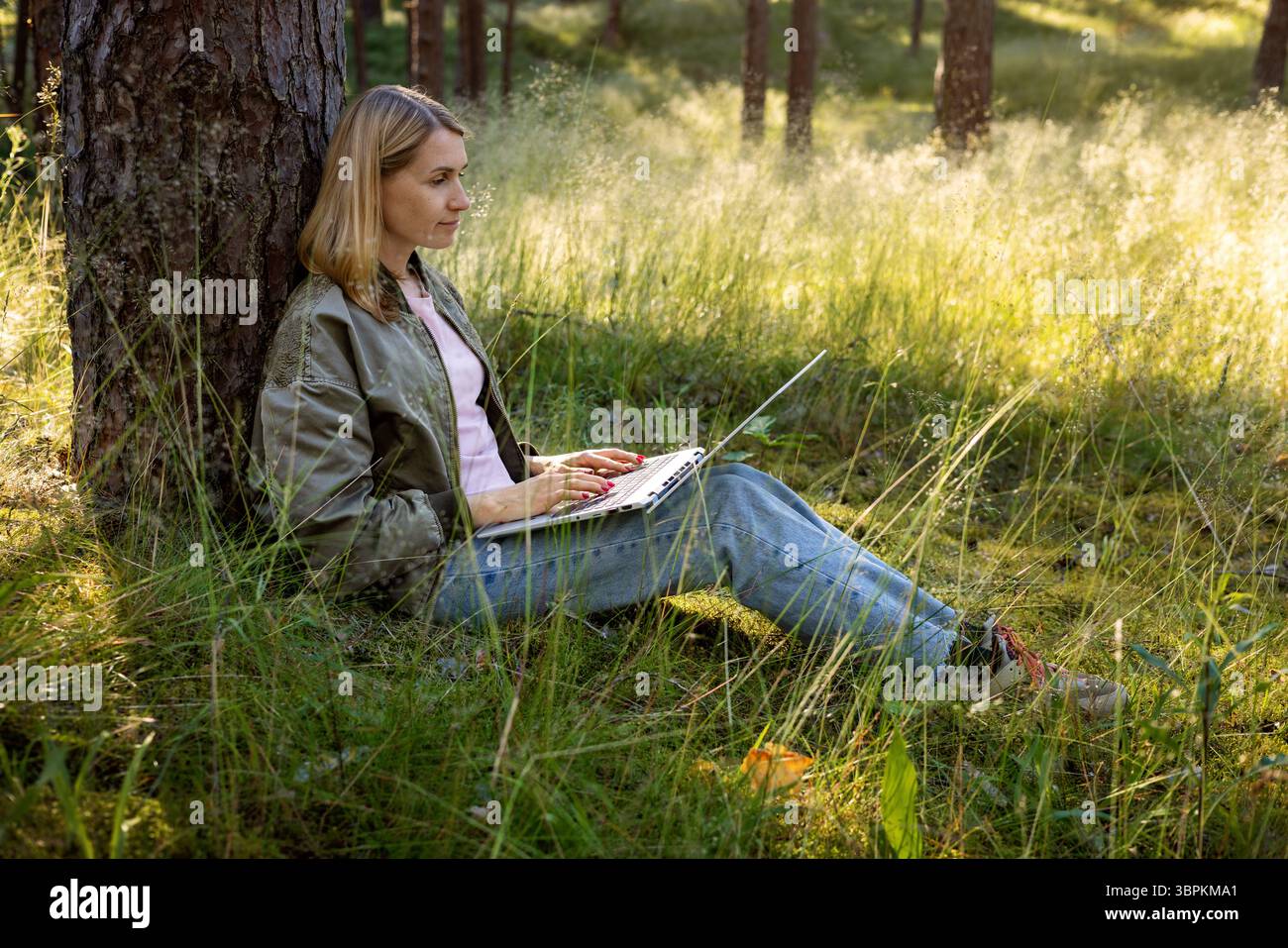 lavoratrice autonoma che utilizza un notebook nel parco. nomade digitale, proprietario di una piccola impresa seduto contro l'albero e lavora in remoto in natura. remo freelance Foto Stock