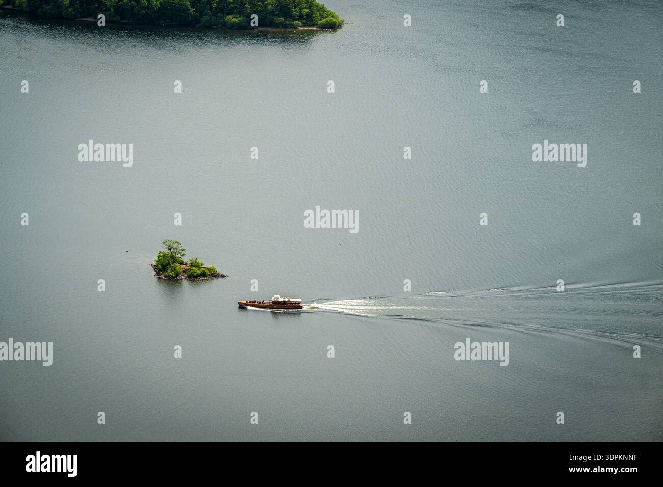 Vista aerea di un traghetto che vola sulla tranquilla superficie del lago Derwentwater, passando vicino a una piccola isola e lasciandosi alle spalle un dolce sentiero d'acqua Foto Stock