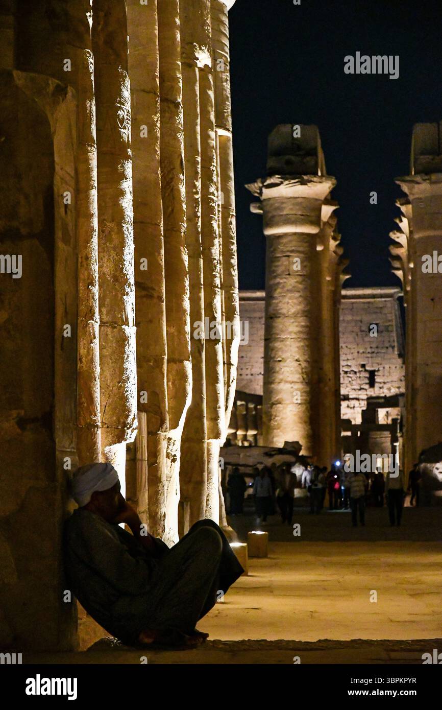 Scena notturna al Tempio di Luxor con un uomo egiziano seduto all'ombra contro antiche colonne illuminate, Luxor, Egitto Foto Stock