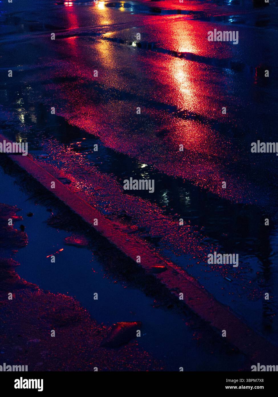 I riflessi notturni danzano sulle pozzanghere d'acqua sul marciapiede, mostrando colori vivaci dai lampioni e dagli edifici circostanti in un ambiente urbano Foto Stock