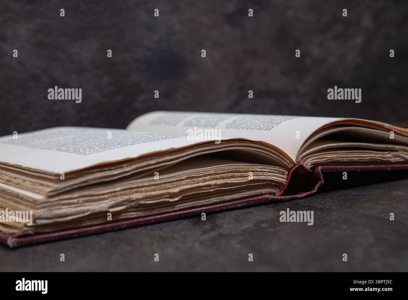 Chiudere la vista laterale di un libro antico aperto, concentrarsi sui bordi della pagina sganciati, poggiare su una superficie a trama scura con sfondo morbido e prospettiva angolata Foto Stock