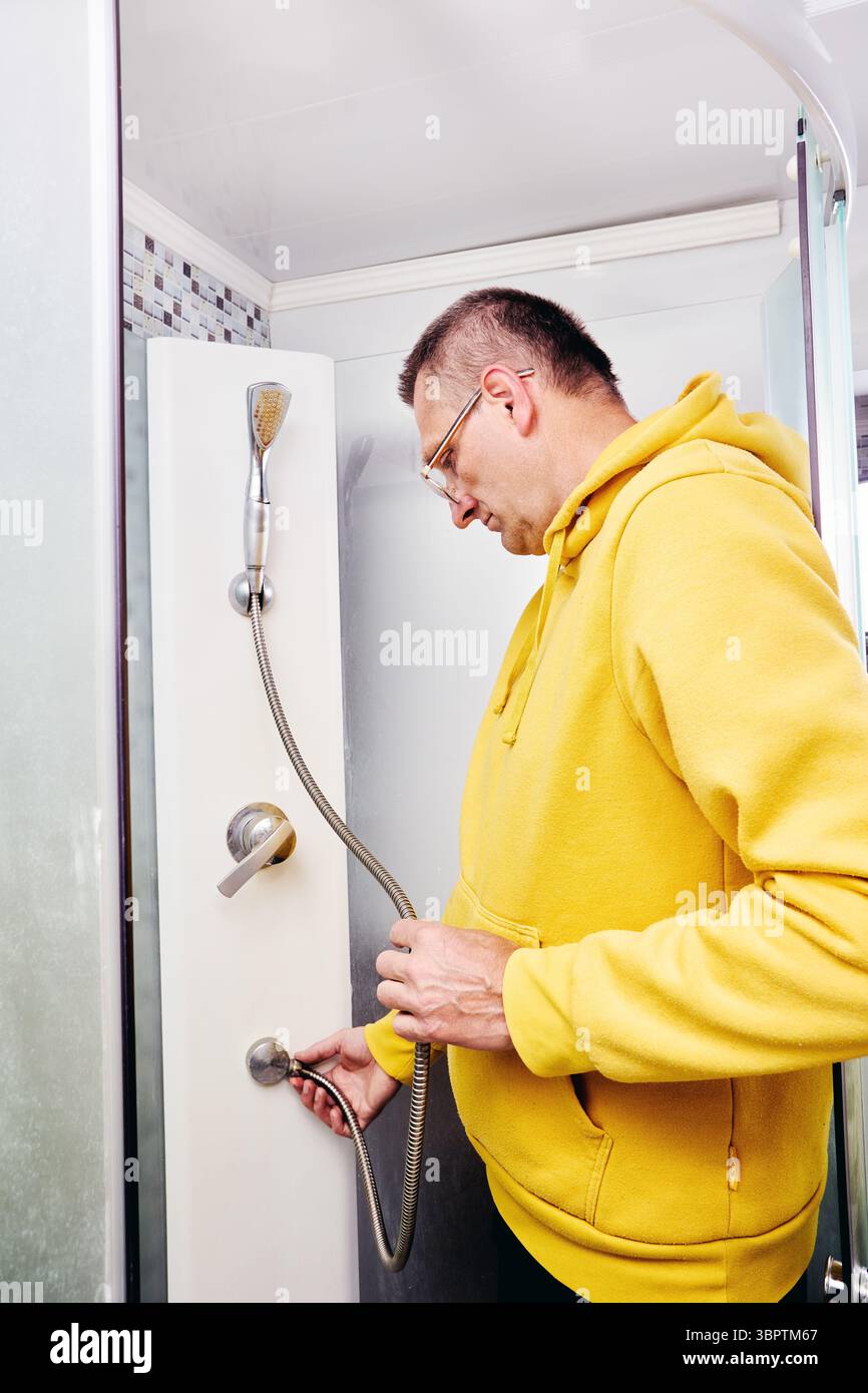 L'uomo all'interno della cabina doccia ispeziona e stringe il raccordo del tubo della doccia sul pannello inferiore per riparare le perdite nel bagno moderno. Foto Stock