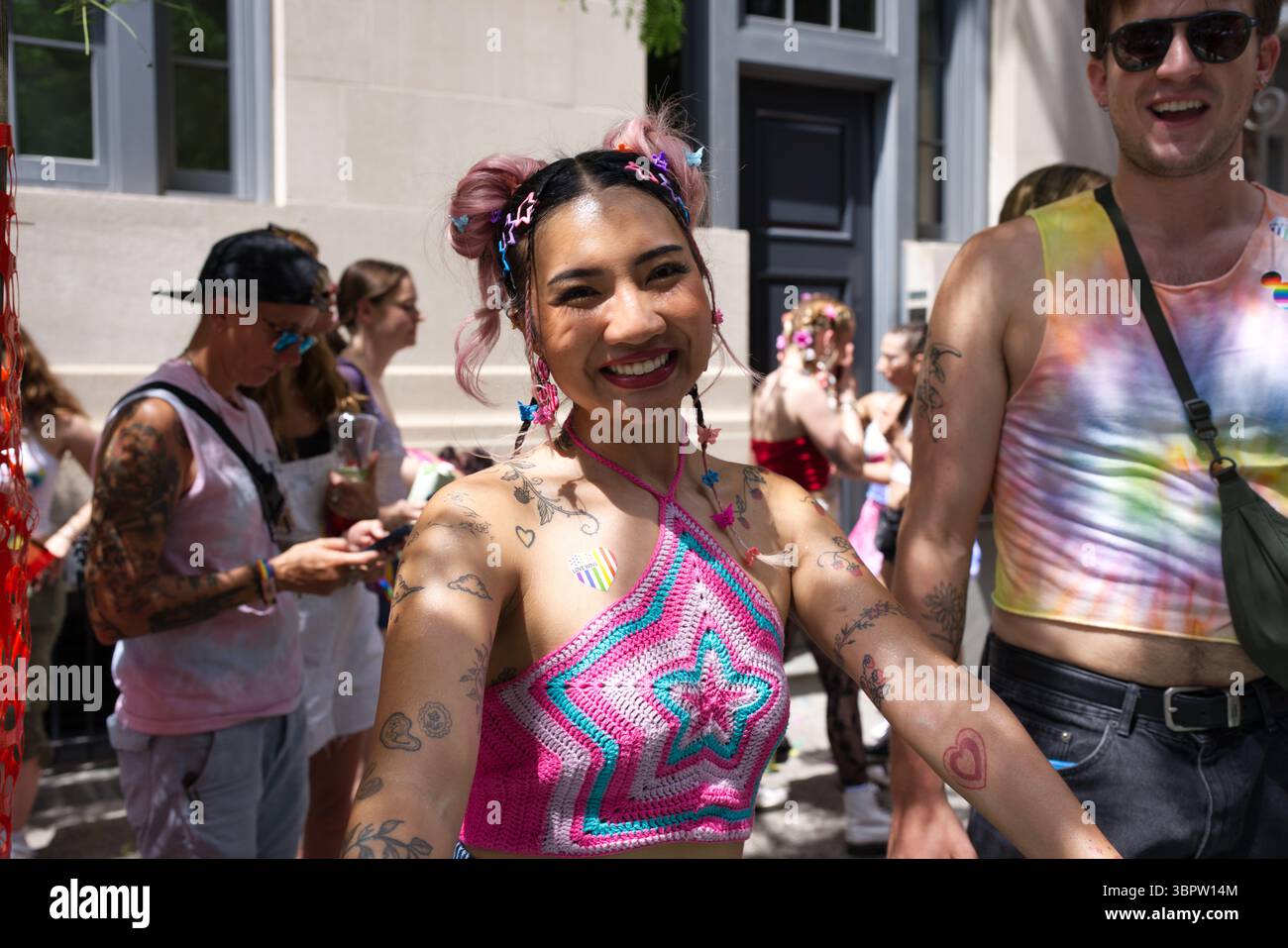 Una giovane donna con i capelli rosa e numerosi tatuaggi sorride alla macchina fotografica, indossando una parte superiore con un collo all'uncinetto. È circondata da altre persone in un outdoo Foto Stock