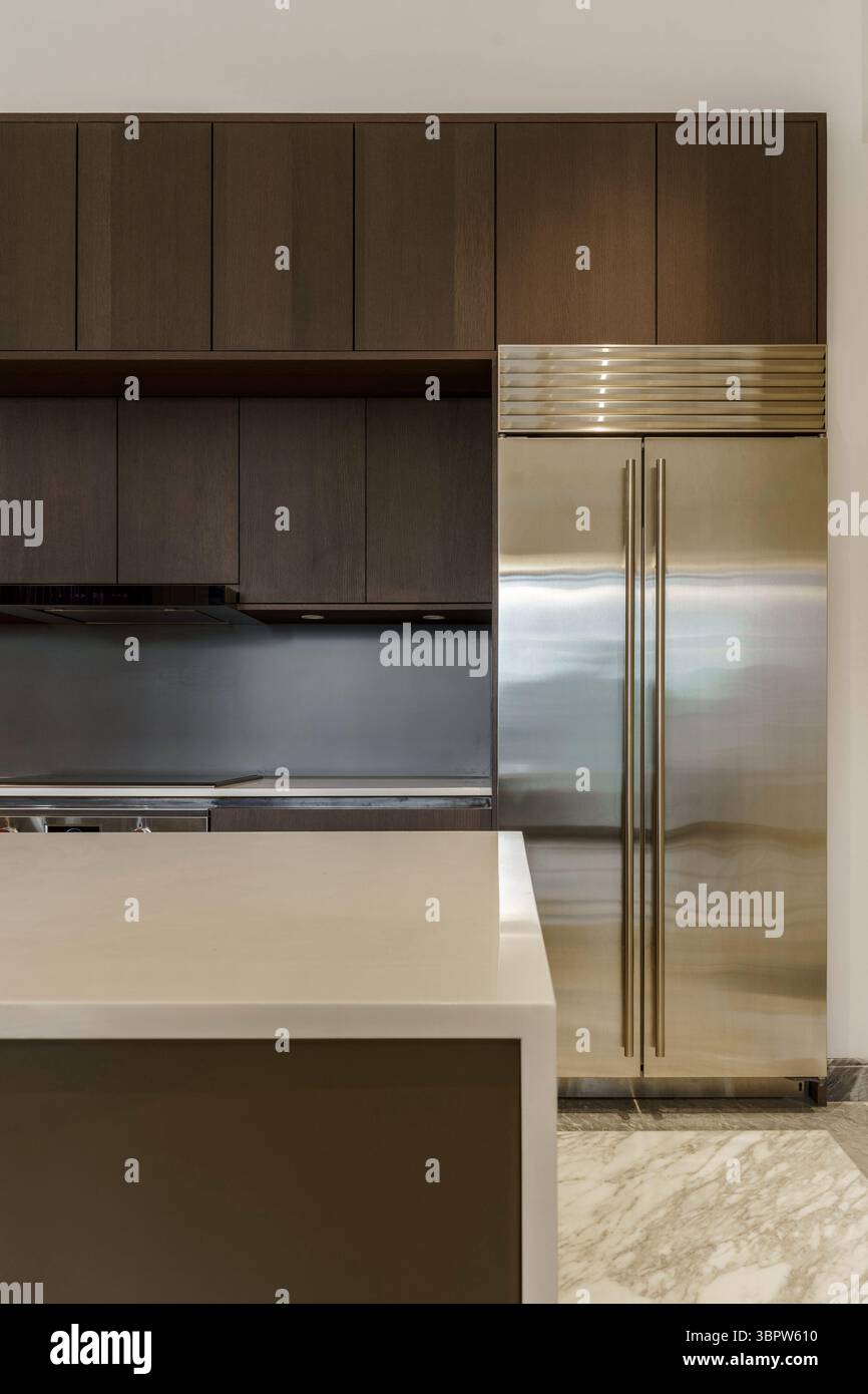 Cucina moderna con un grande frigorifero incorporato in acciaio inox e armadietti in legno scuro. Interni eleganti con un ripiano bianco e pavimento in marmo. Foto Stock
