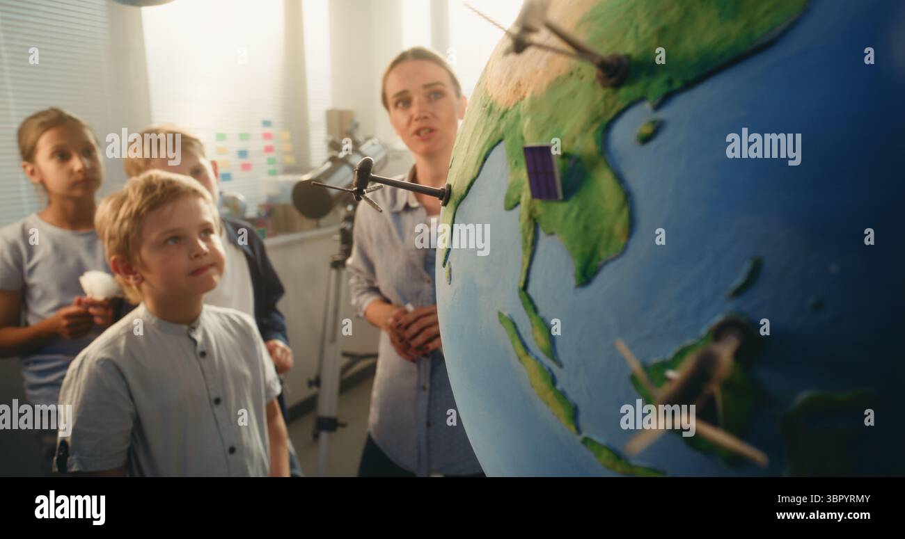 Classe Ecologia STEM: Scuola elementare ragazzo che attacca il modello in miniatura della turbina eolica a Globe. Insegnante donna che spiega la scienza e le fonti di energia rinnovabile agli studenti intelligenti in classe moderna. Foto Stock