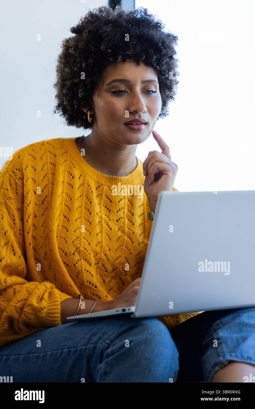 Donna che indossa un maglione giallo senape che lavora con cura su un notebook argentato a casa. Produttività, comfort, moderno, informale, spazio di lavoro, concentrati, tecnologia Foto Stock