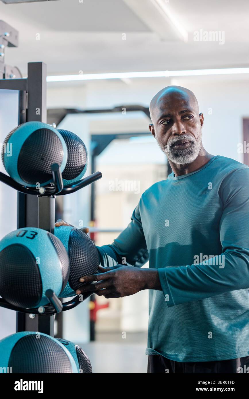 Uomo afroamericano senior che seleziona palla medica da un rack con una camicia di tè nell'area palestra. Fitness, forza, salute, determinazione, allenamento, sforzo Foto Stock