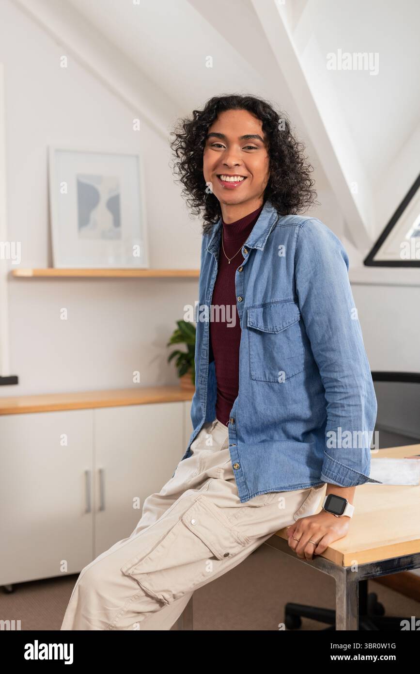Persona non binaria appoggiata su una scrivania in legno in un ufficio in stile loft con stampa astratta incorniciata. Contemporaneo, minimalista, spazio di lavoro, creativo, naturale Foto Stock
