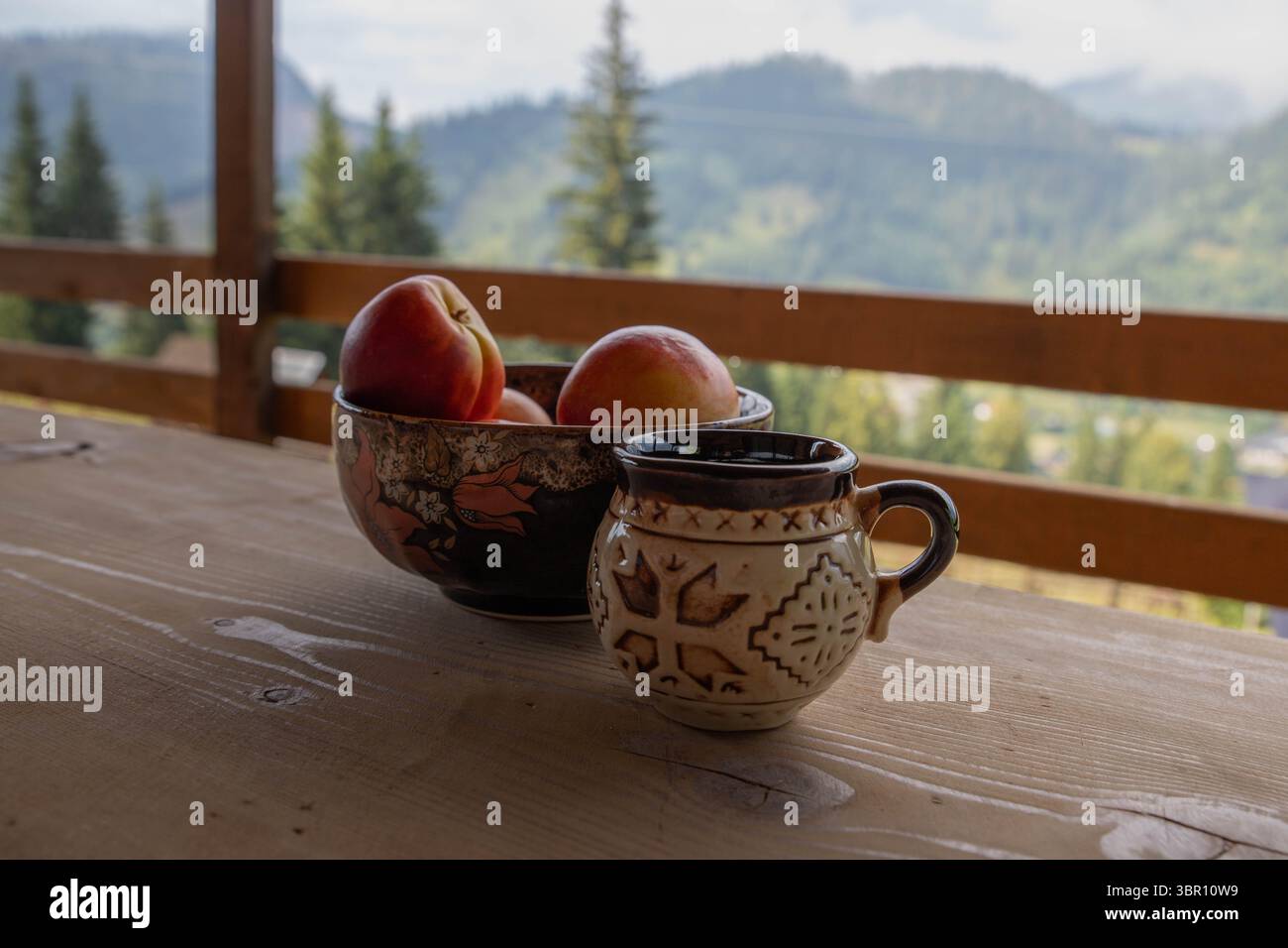 pesche rosse e profumate in una ciotola di argilla smaltata e una tazza di argilla smaltata su un rustico tavolo di legno naturale, sfondo con paesaggio montano Foto Stock