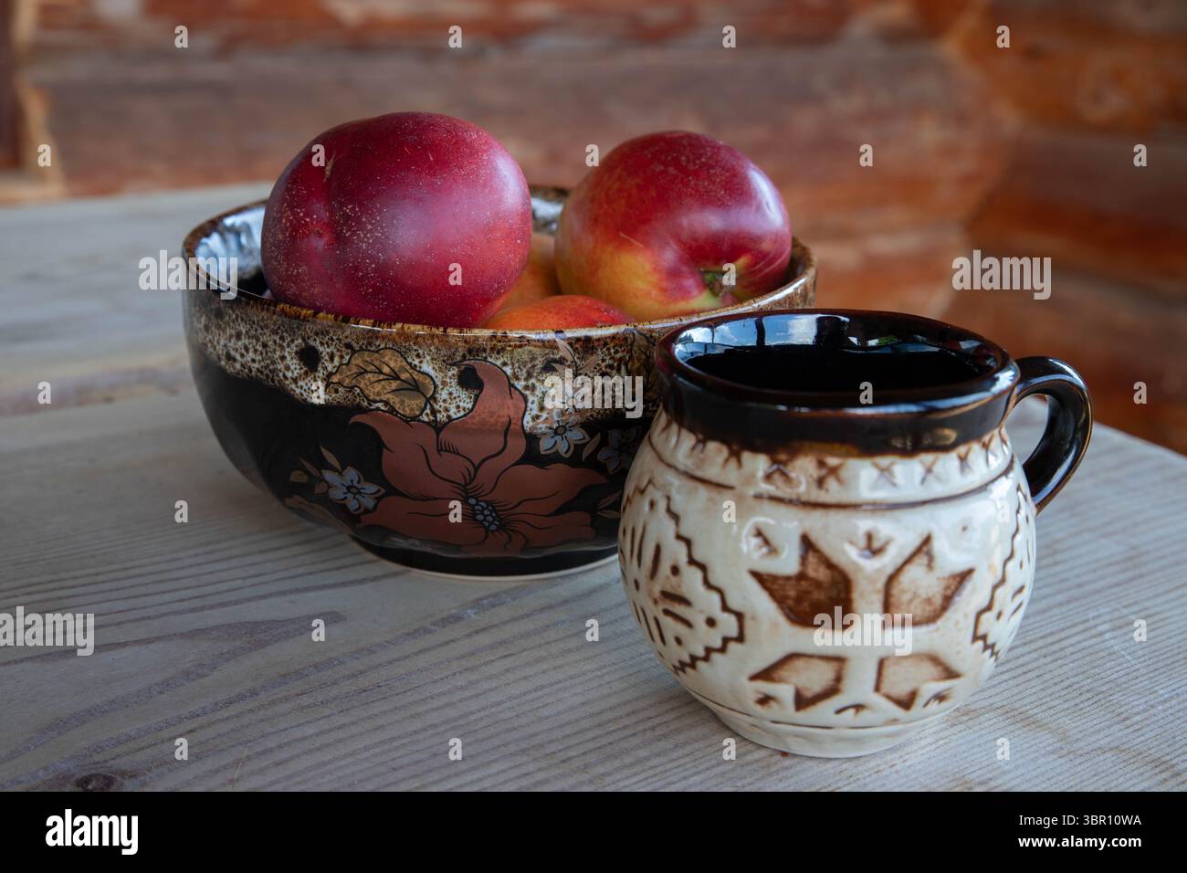 pesche rosse e profumate in una ciotola di argilla smaltata e una tazza di argilla smaltata su un rustico tavolo in legno naturale, sfondo con parete in legno della cabina Foto Stock