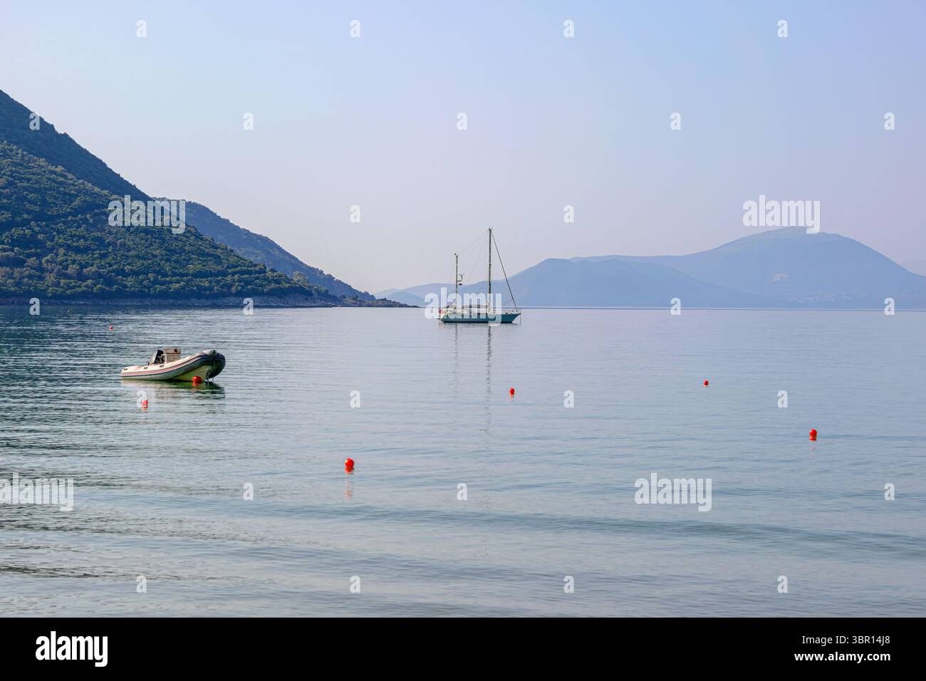 Una barca a vela e un gommone ancorati nelle calme acque riflettenti di una baia, con montagne nebbiose sullo sfondo sotto un cielo morbido a Vasiliki Beach. Foto Stock