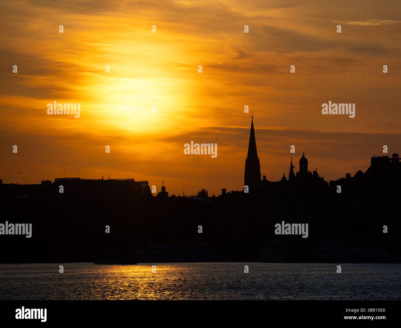 Uno splendido tramonto sullo skyline della città, sagome di edifici e una guglia prominente contro un vivace cielo arancione e giallo. L'acqua riflette il Foto Stock