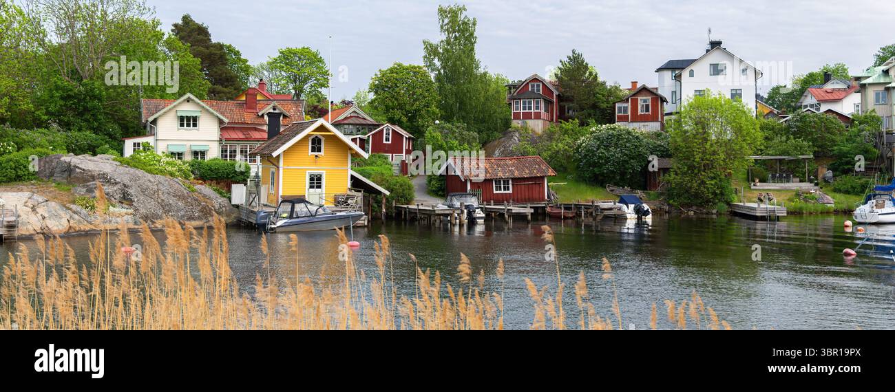Una pittoresca scena costiera sull'isola di Vaxholm caratterizzata da case di legno colorate lungo un tranquillo corso d'acqua. Le barche sono attraccate ai moli, circondati da l Foto Stock