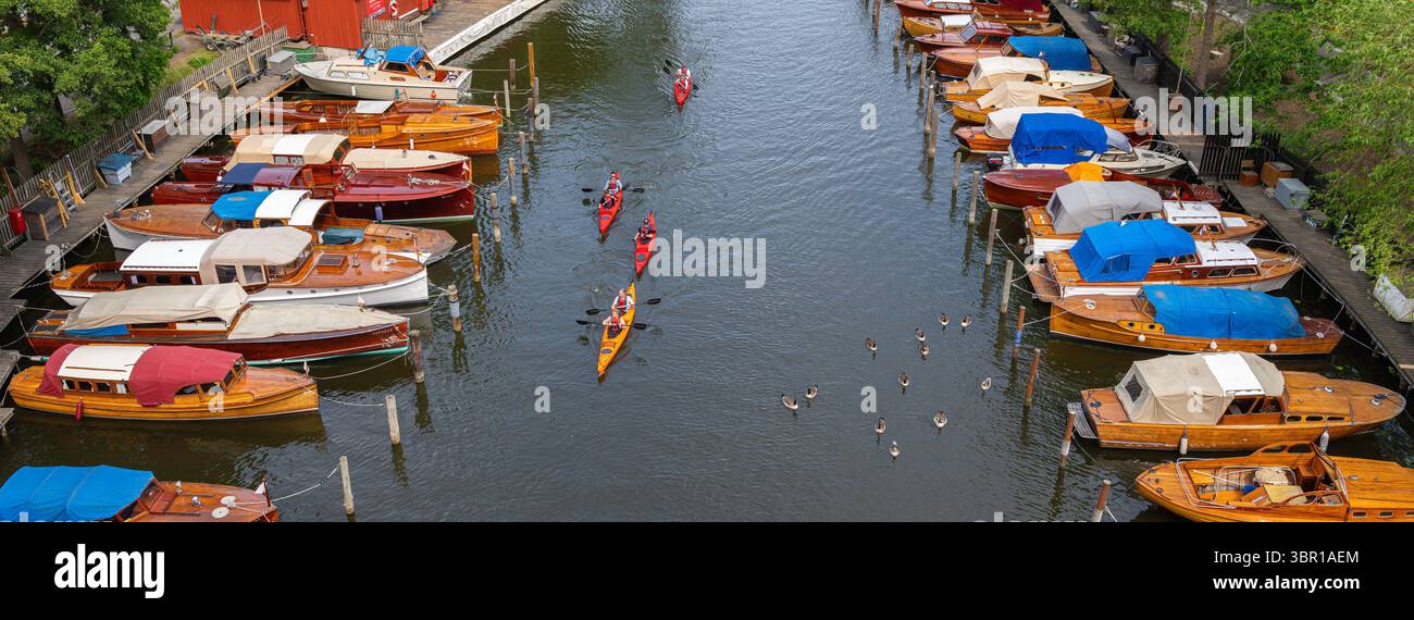 Stoccolma, Svezia - 10 giugno 2025: Veduta aerea di un tranquillo corso d'acqua costeggiato da barche in legno, alcune ricoperte di teloni blu. I kayak navigano attraverso t Foto Stock