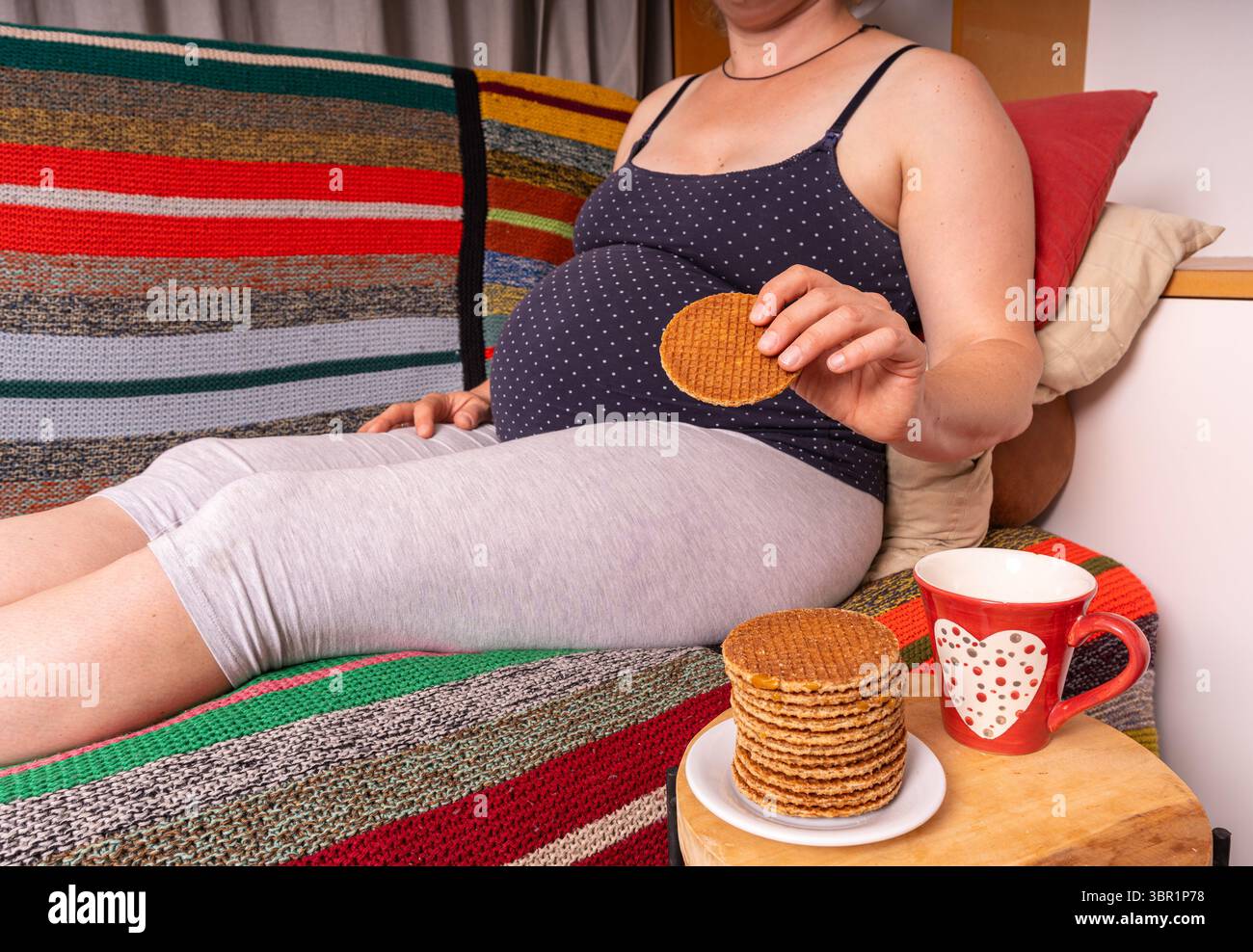 Donna incinta seduta sul divano di casa che si gusta biscotti, momento di maternità rilassato Foto Stock