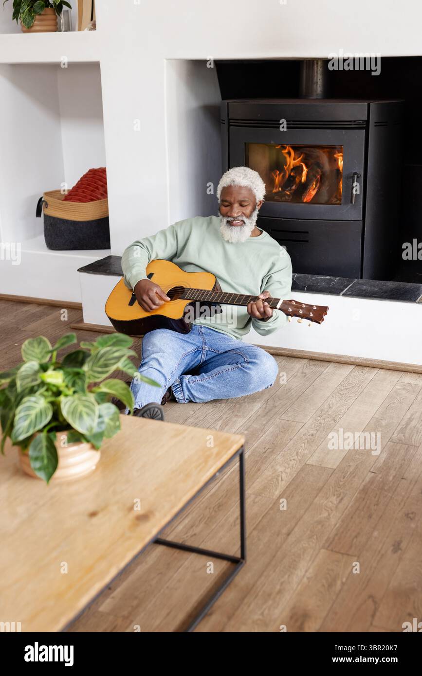 Uomo afroamericano senior che suona la chitarra nel soggiorno con tavolino da caffè, pianta e fornello. Musicista, relax, accogliente, campagna, acustica, no Foto Stock