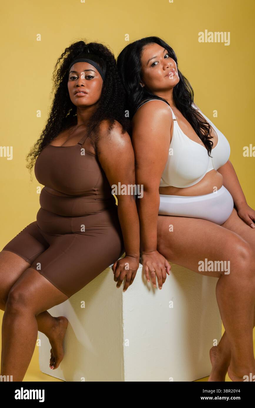 Diverse amiche in studio sedute dietro a un cubo bianco che sfoggia un look di forma. Positività corporea, fiducia, diversità, empowerment, moda Foto Stock