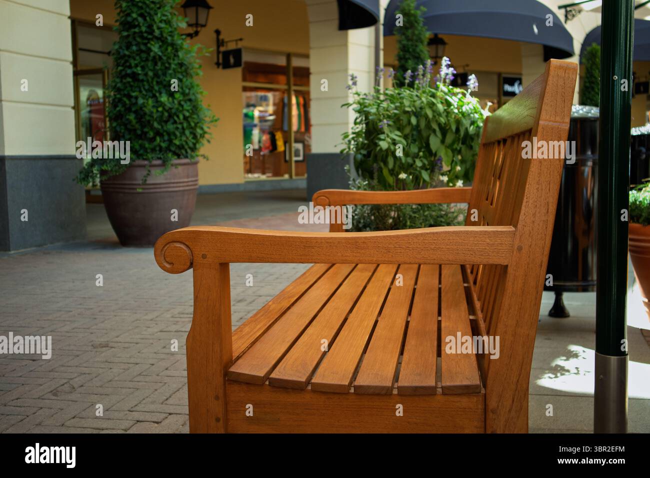 Una panca in legno classica e robusta si stende vuota su un passaggio pavimentato, invitando a riposarsi all'esterno dei negozi all'interno di un luminoso ambiente commerciale all'aperto Foto Stock