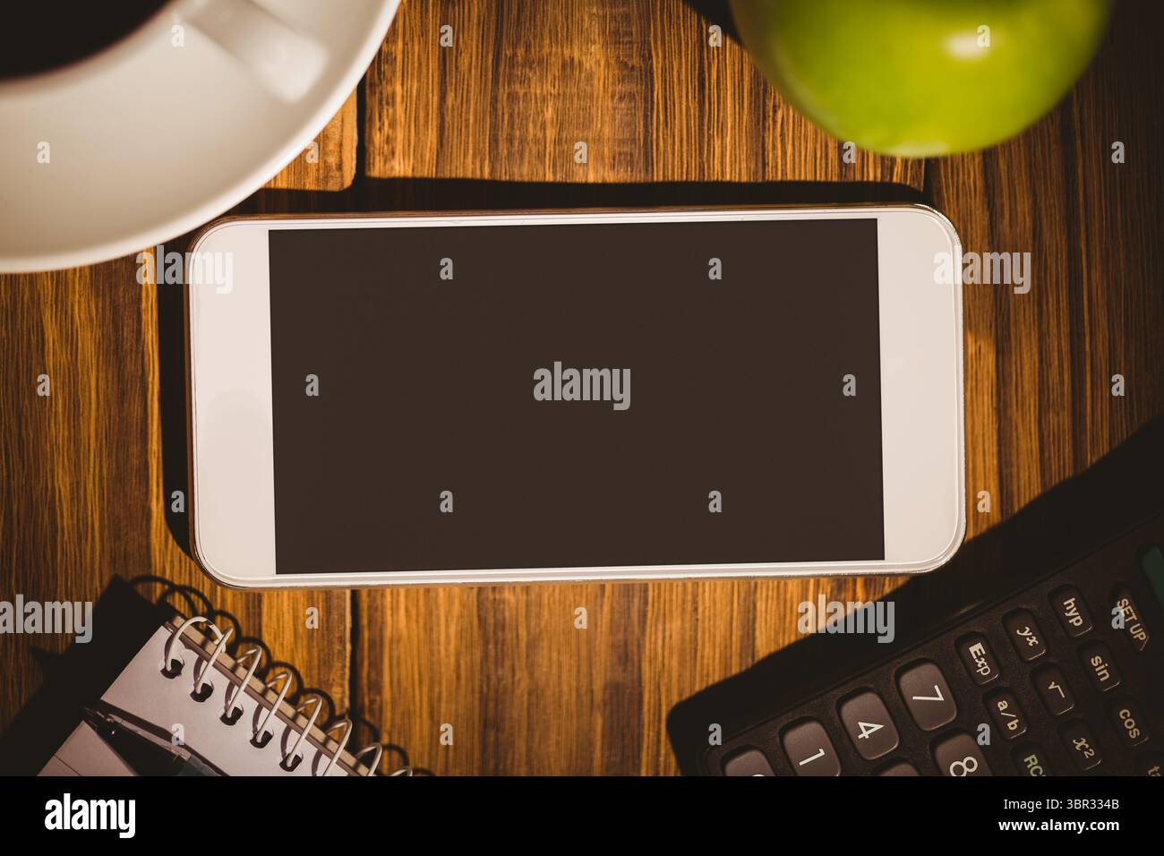 Lo smartphone con cornice bianca si trova su una scrivania in legno con tazza di caffè, mela verde, notebook e calcolatrice Foto Stock