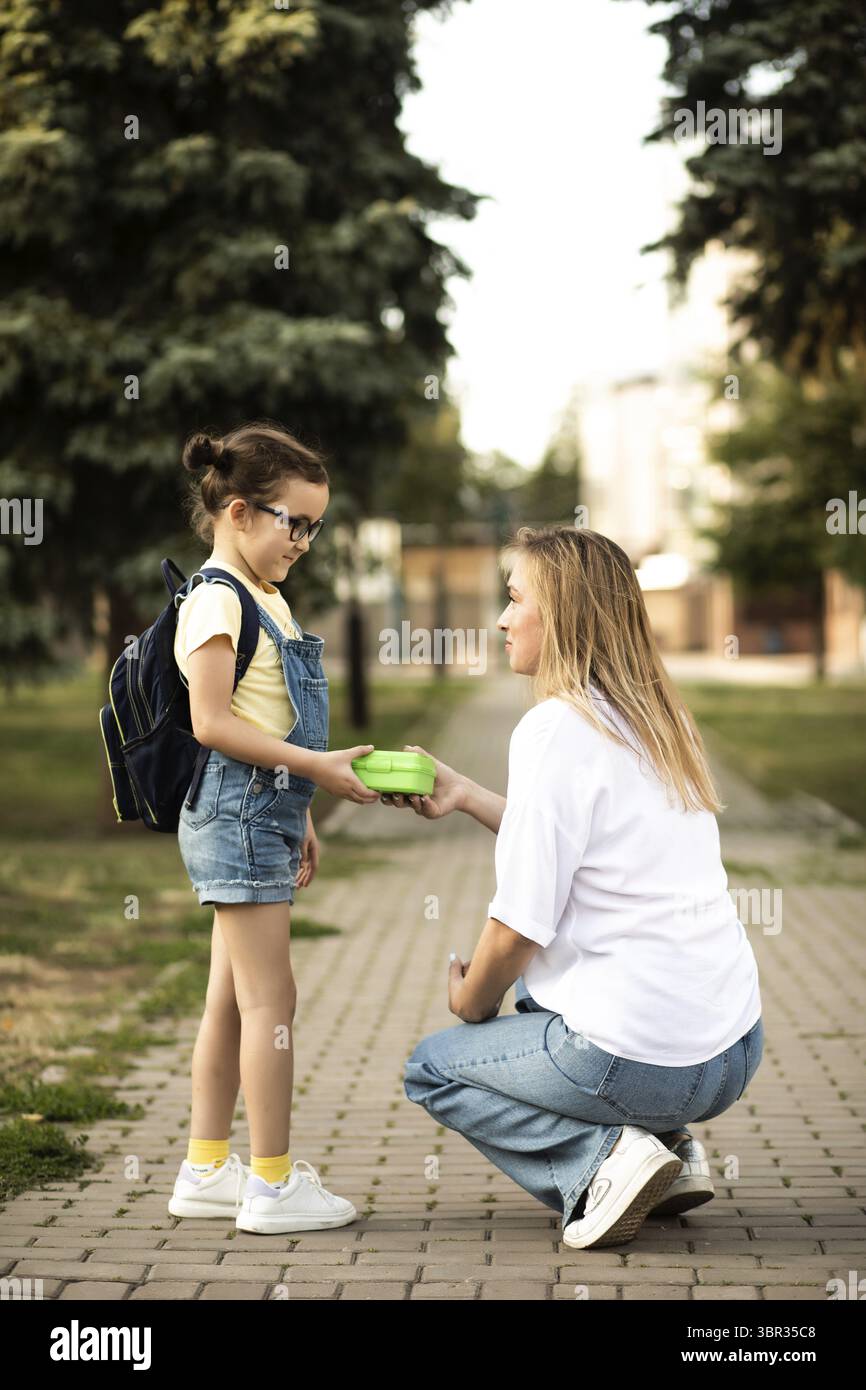Mamma Gibt Tochter niedlichen kleinen Schulmaedchen mit Rucksack Lunch-Box. In der naehe der Schule im Freien. Essen, sempre più gentile Foto Stock