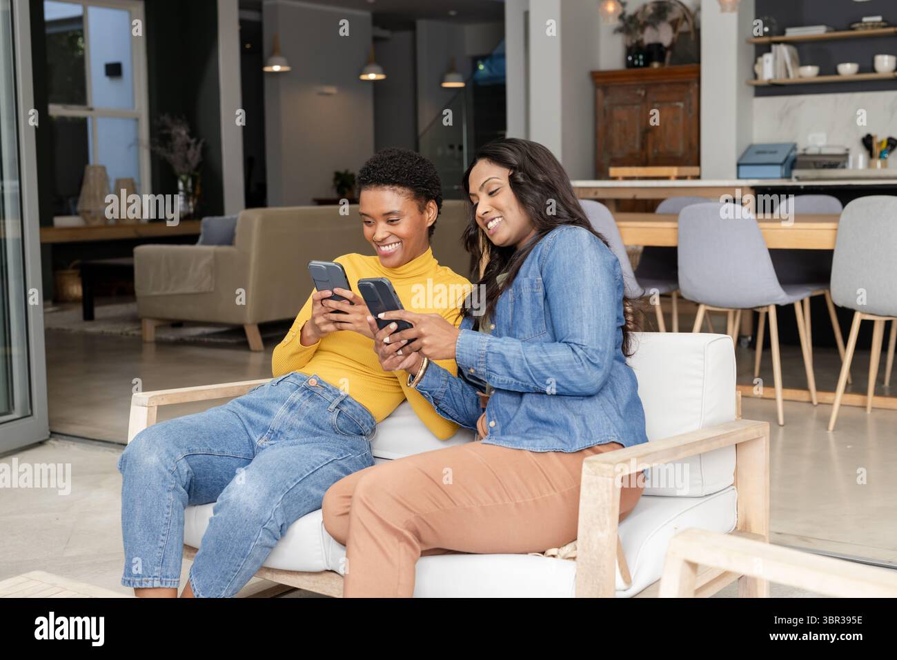 Diverse amiche sedute su un divano imbottito che tiene gli smartphone al tavolo da pranzo in una casa open space Foto Stock