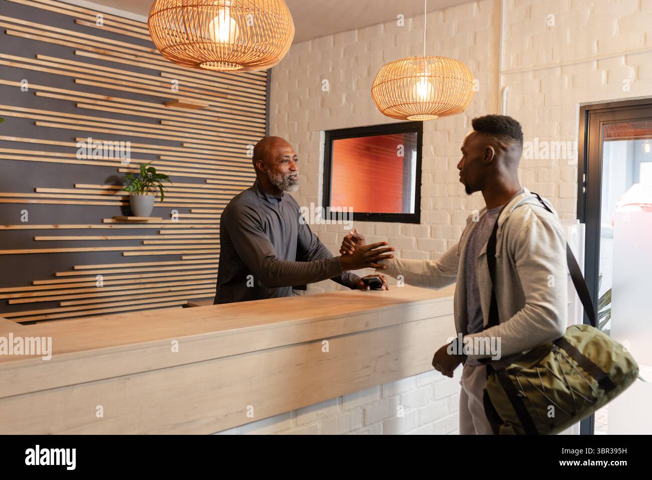 Il receptionist afroamericano e il visitatore stringono la mano al banco della reception della palestra portando con sé il borsone Foto Stock